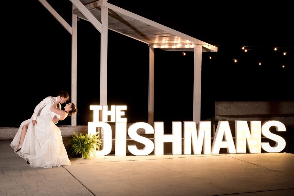 Newlyweds kiss beside illuminated marquee letters spelling THE DISHMANS at their outdoor wedding reception.