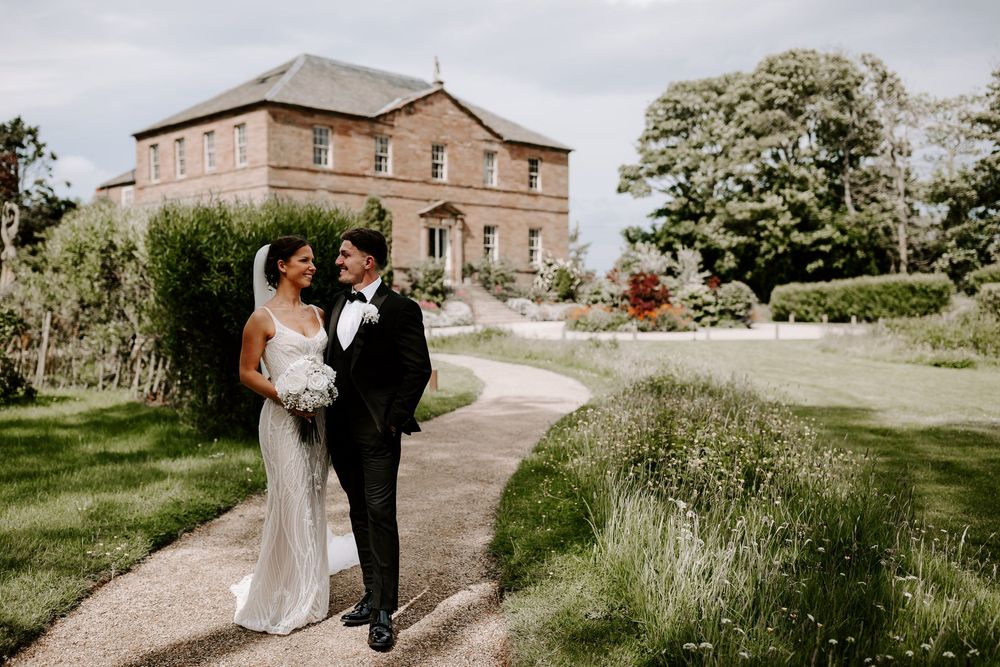 Bride and groom smiling together on a garden path outside a grand stone country house wedding venue.
