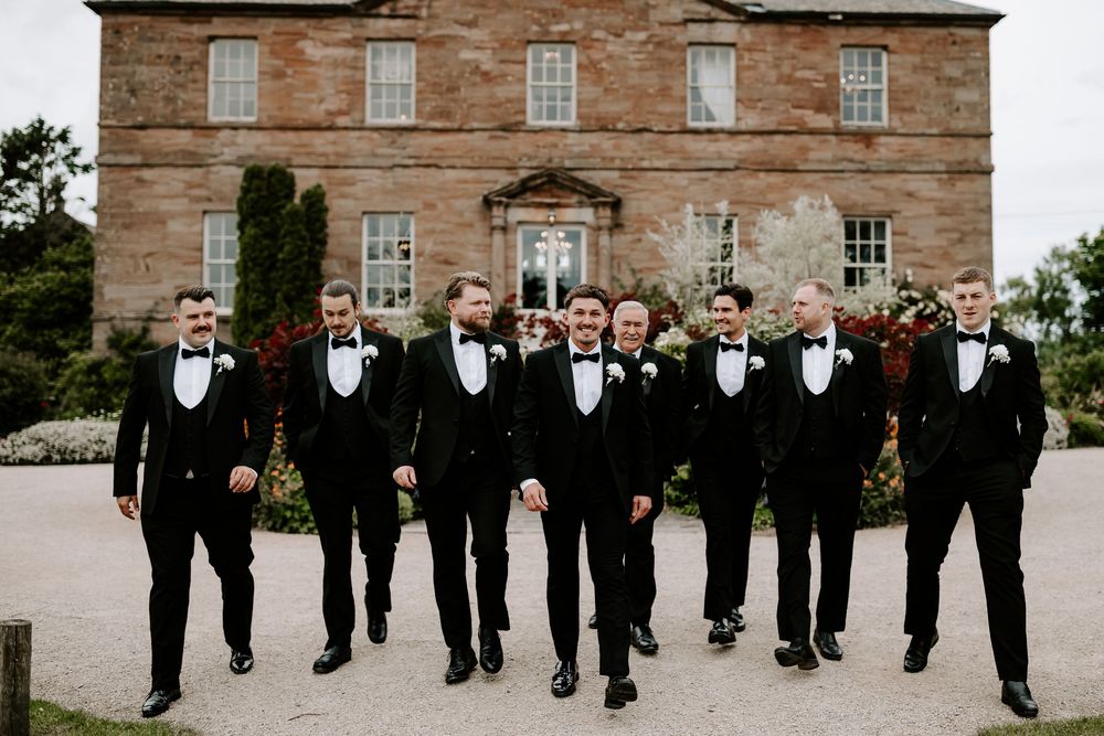 Eight groomsmen in black tuxedos with bow ties walk confidently in front of a historic stone manor house.