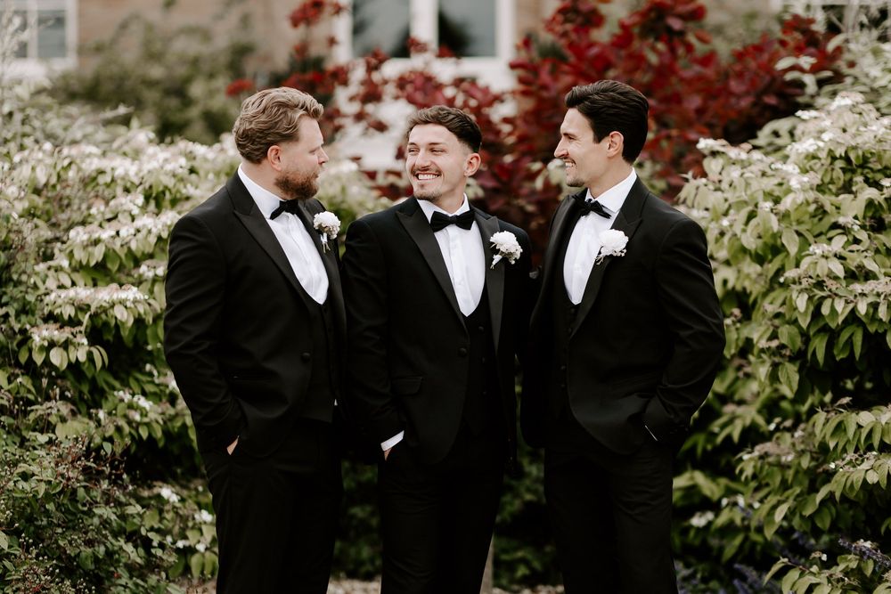 Three groomsmen in black tuxedos with bow ties and boutonnieres laughing together outdoors among lush green and white foliage.