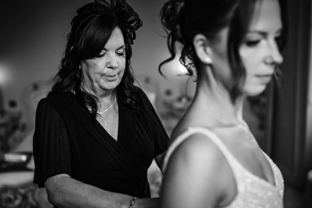 Black and white photo of mother helping bride with wedding dress during bridal preparations.