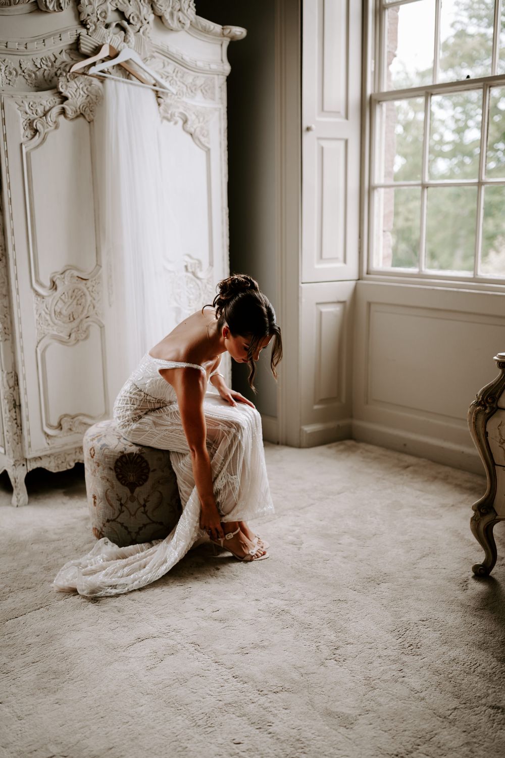 Bride in lace wedding dress sitting on ottoman adjusting her heels near ornate white armoire and window.