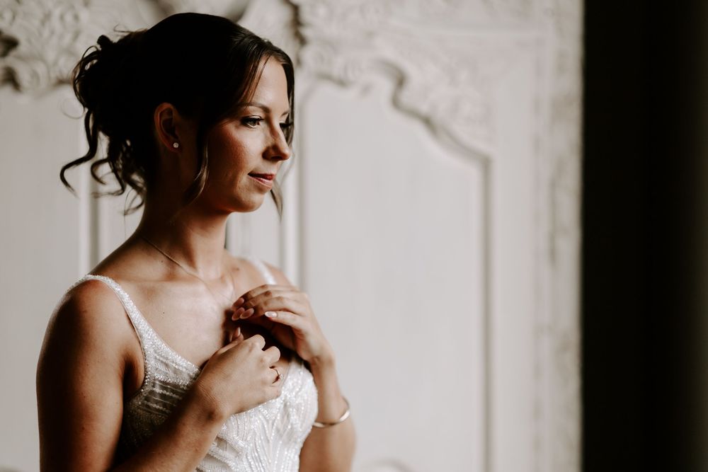 Elegant bride in white lace dress holding jewelry, gazing thoughtfully in soft natural light near ornate wall.