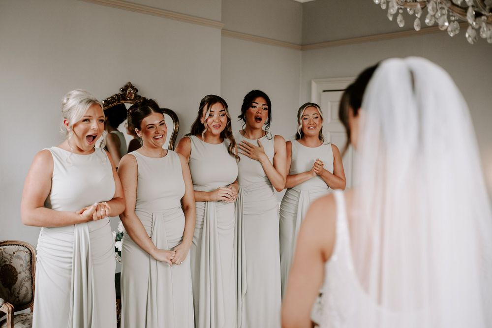 Bridesmaids in sage green gowns react emotionally to bride in white wedding dress and veil during first look.