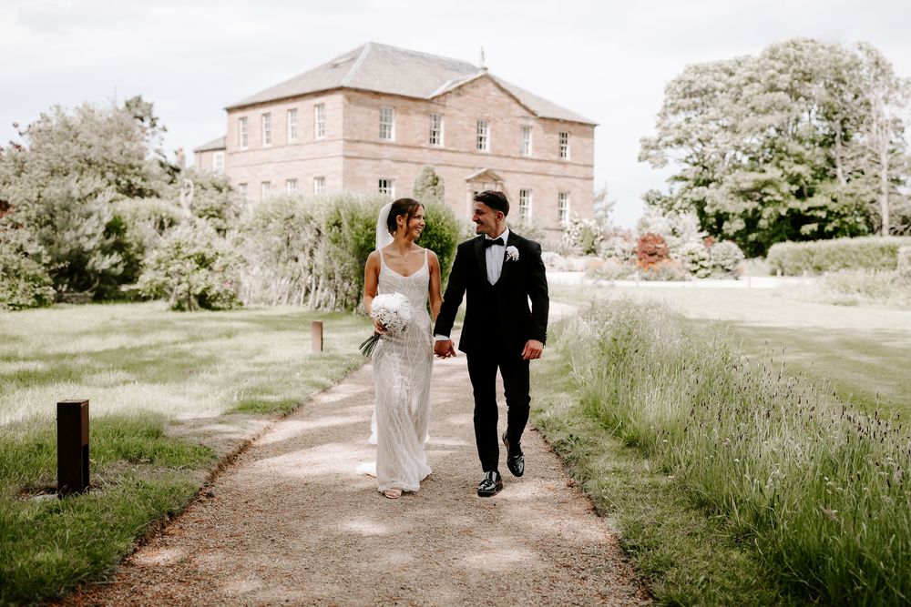 Bride and groom walk hand in hand along a garden path toward an elegant stone manor on their wedding day.