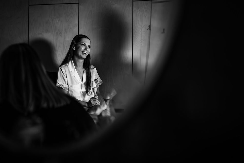Black and white photo of a smiling bride in a white robe during wedding preparations, captured through a mirror reflection.