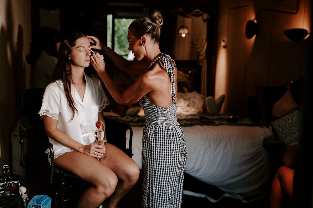 A makeup artist in a gingham dress applies makeup to a seated woman in a warmly lit rustic room.