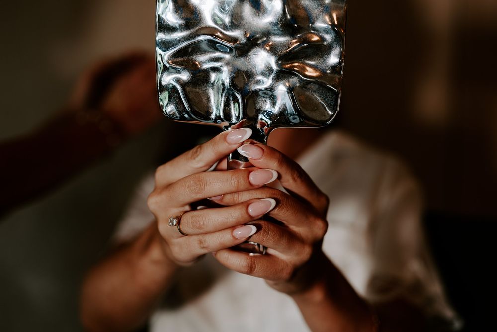 Hands with pink manicure and rings holding a crinkled silver metallic square mirror.