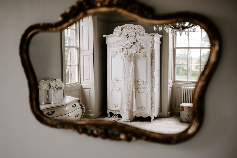 Ornate gold mirror reflecting a white baroque armoire with wedding dress in an elegant vintage room.