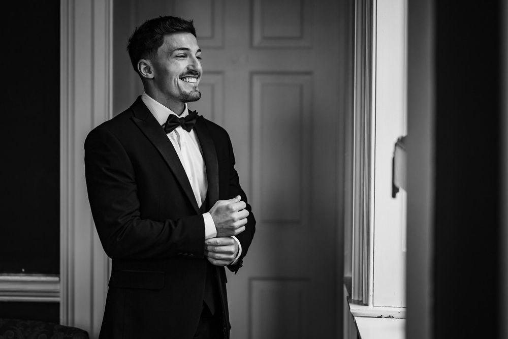 Black and white photo of a smiling groom in a tuxedo adjusting his cufflinks near a window.