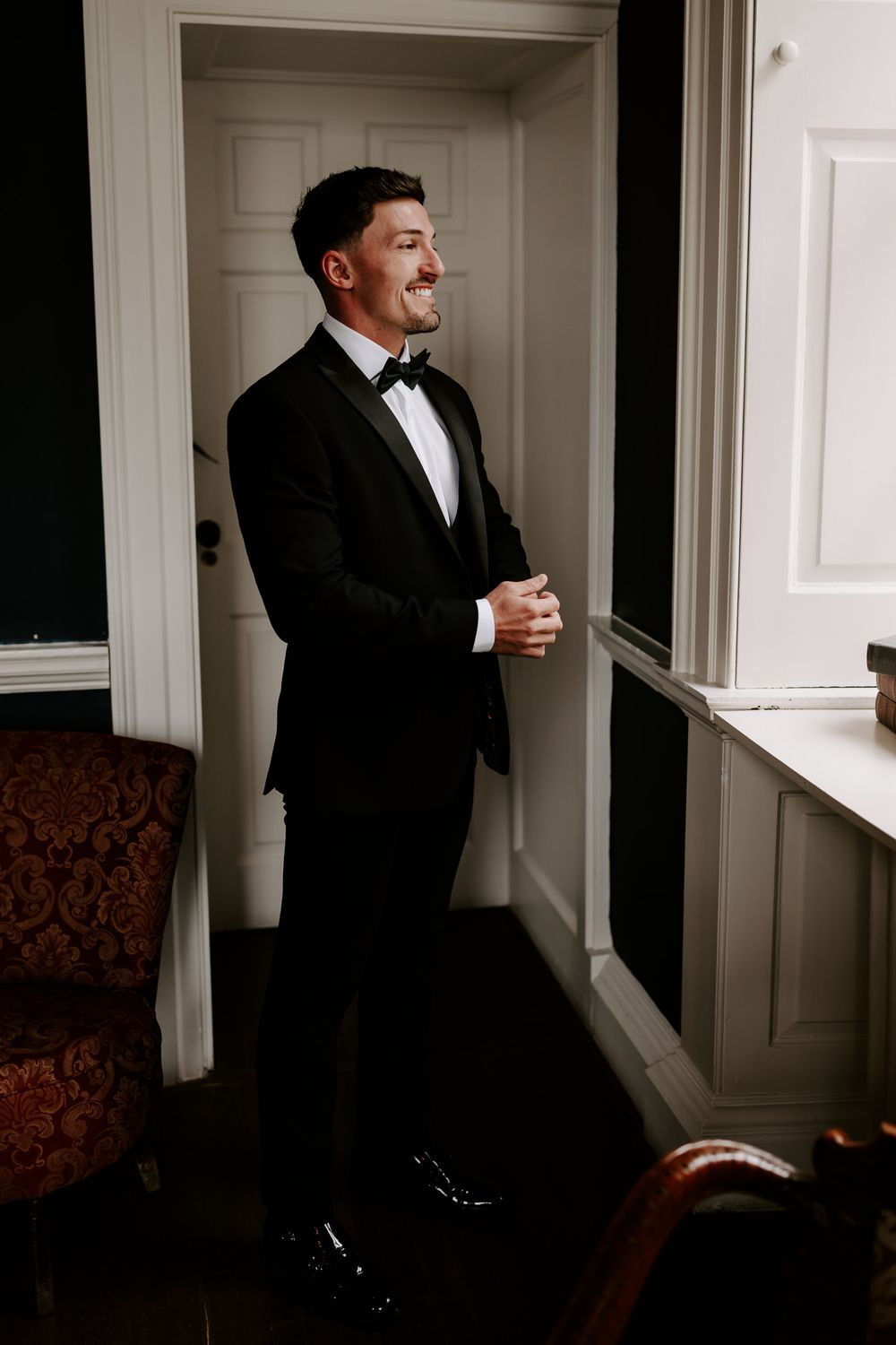 A groom in a black tuxedo and bow tie stands by a window, smiling with hands clasped in an elegant room.