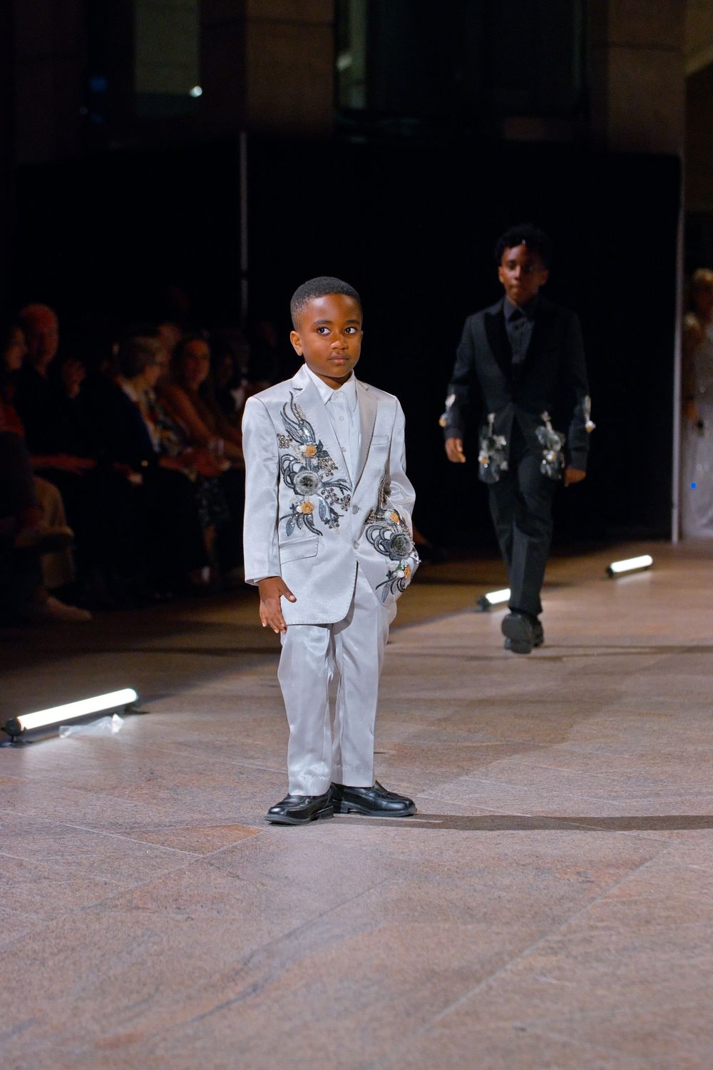 Young boy model in silver embroidered suit walks runway at children's fashion show with audience seated behind.