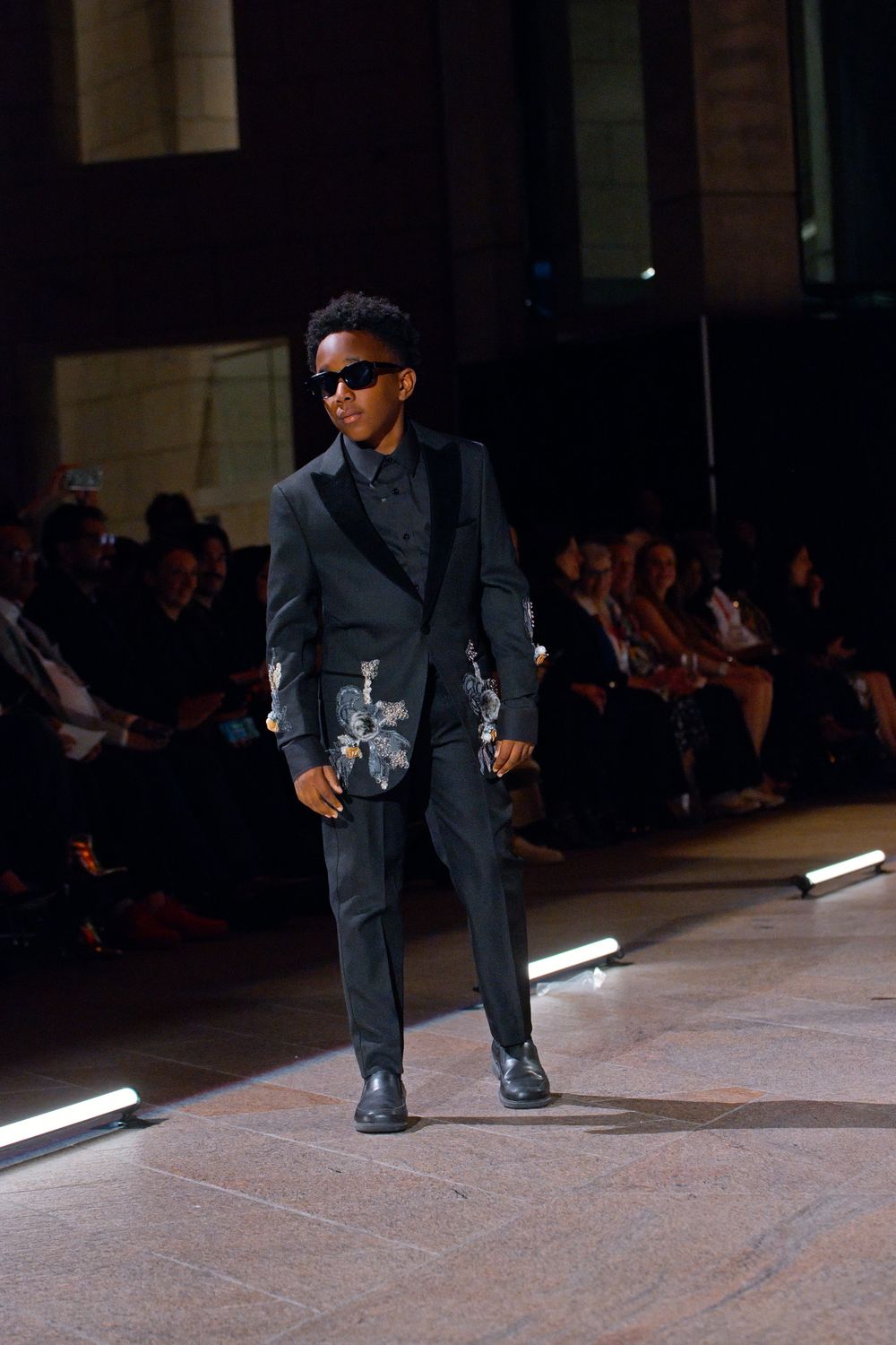 Young male model walks runway in dark floral embroidered suit with sunglasses at fashion show.