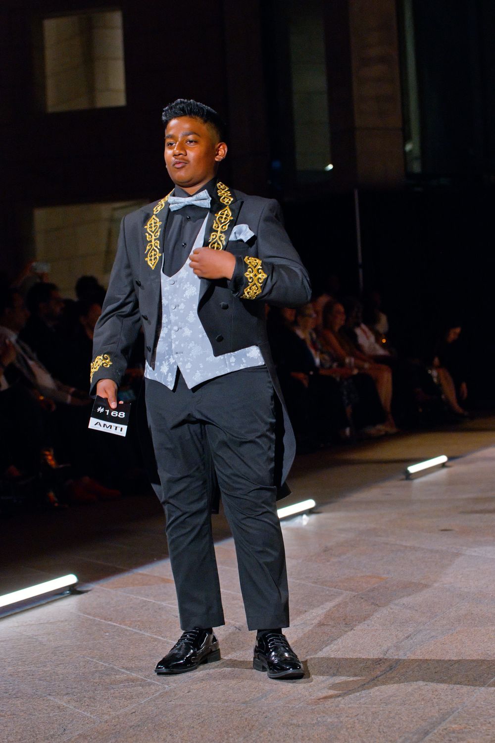 Young male model walks runway in ornate dark tuxedo with gold embroidery, light blue vest and bow tie at fashion show.