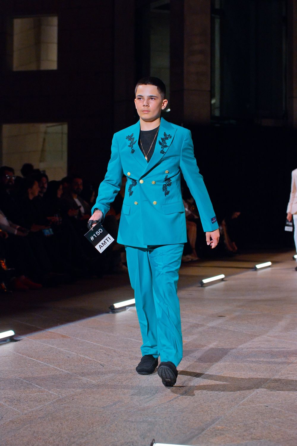 Male model walks runway in turquoise double-breasted suit with black embroidery at fashion show.
