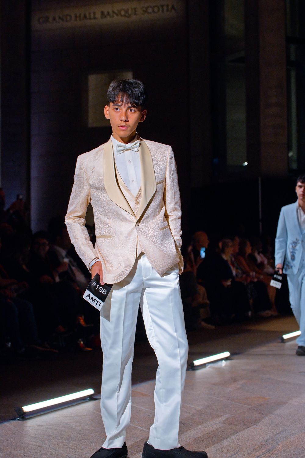 Male model walks runway in white tuxedo with gold lapels and bow tie at Grand Hall Banque Scotia fashion show.