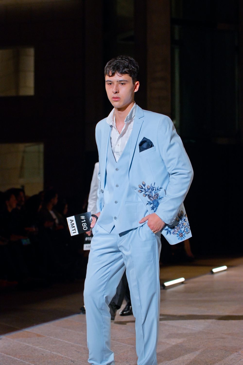 Male model walks runway in light blue three-piece suit with floral embroidery at fashion show.