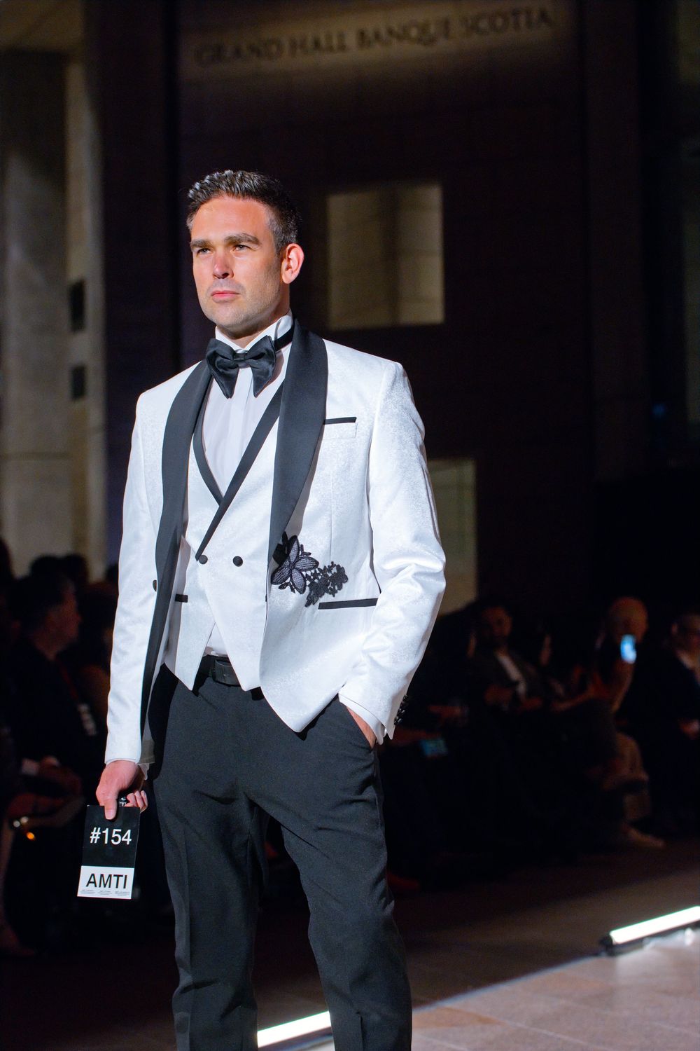 Male model wearing white tuxedo jacket with black lapels and bow tie on runway at Grand Hall Banque Scotia.