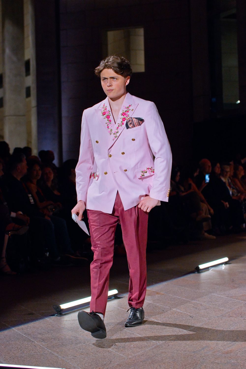 Male model walks runway in pink double-breasted blazer with floral embroidery and rose-colored trousers at fashion show.