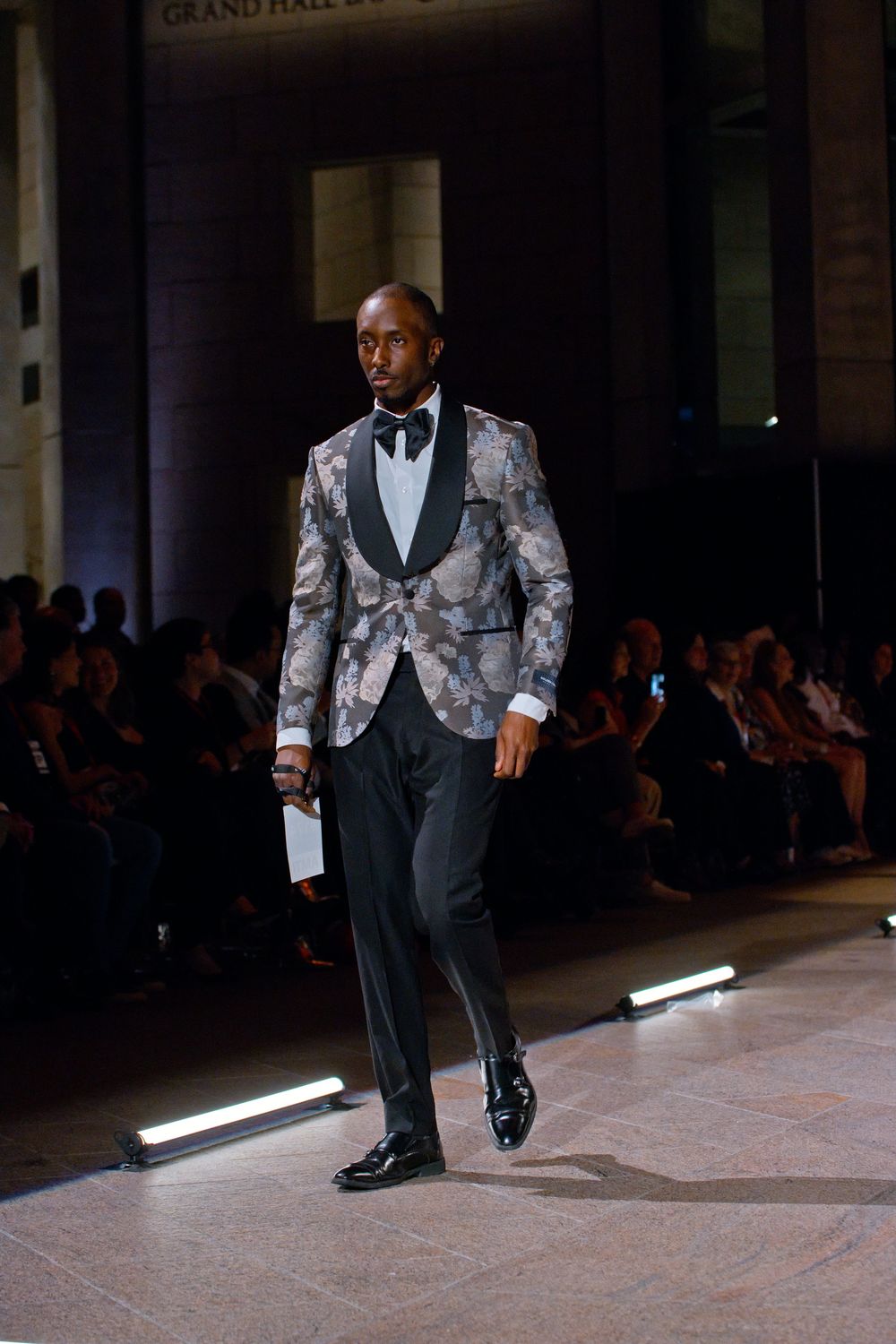 Male model walks runway in floral silver tuxedo jacket with black lapels and trousers at Grand Hall fashion show.