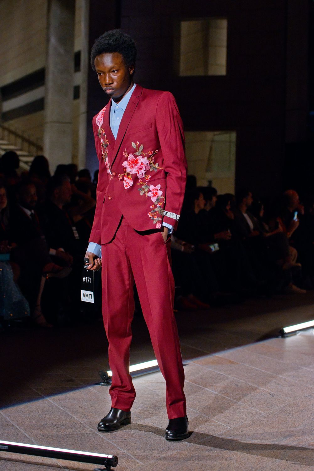 Male model walks runway in a bold red floral embroidered suit with black shoes at a fashion show.