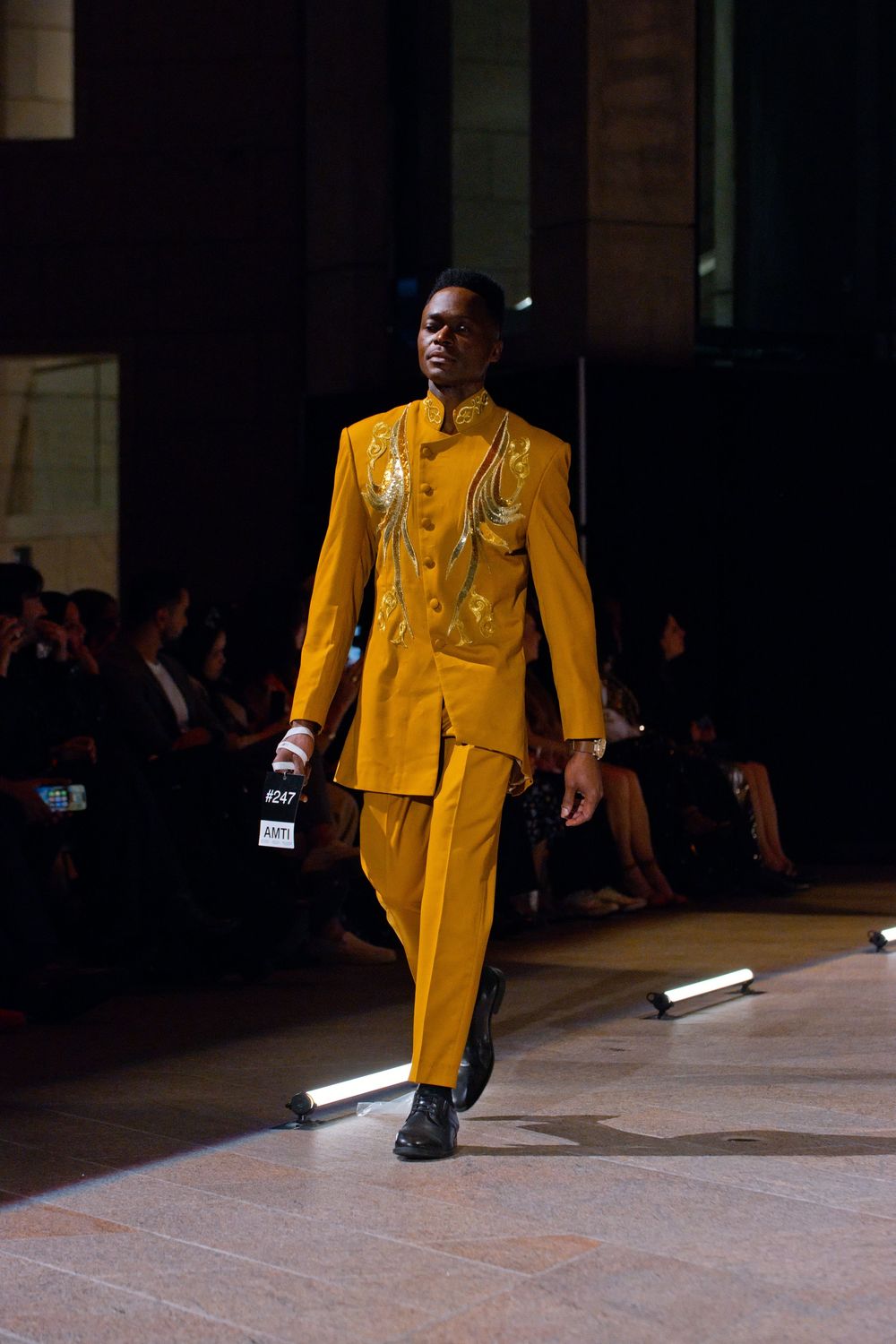 Male model walks runway in gold embroidered mustard yellow suit at fashion show.