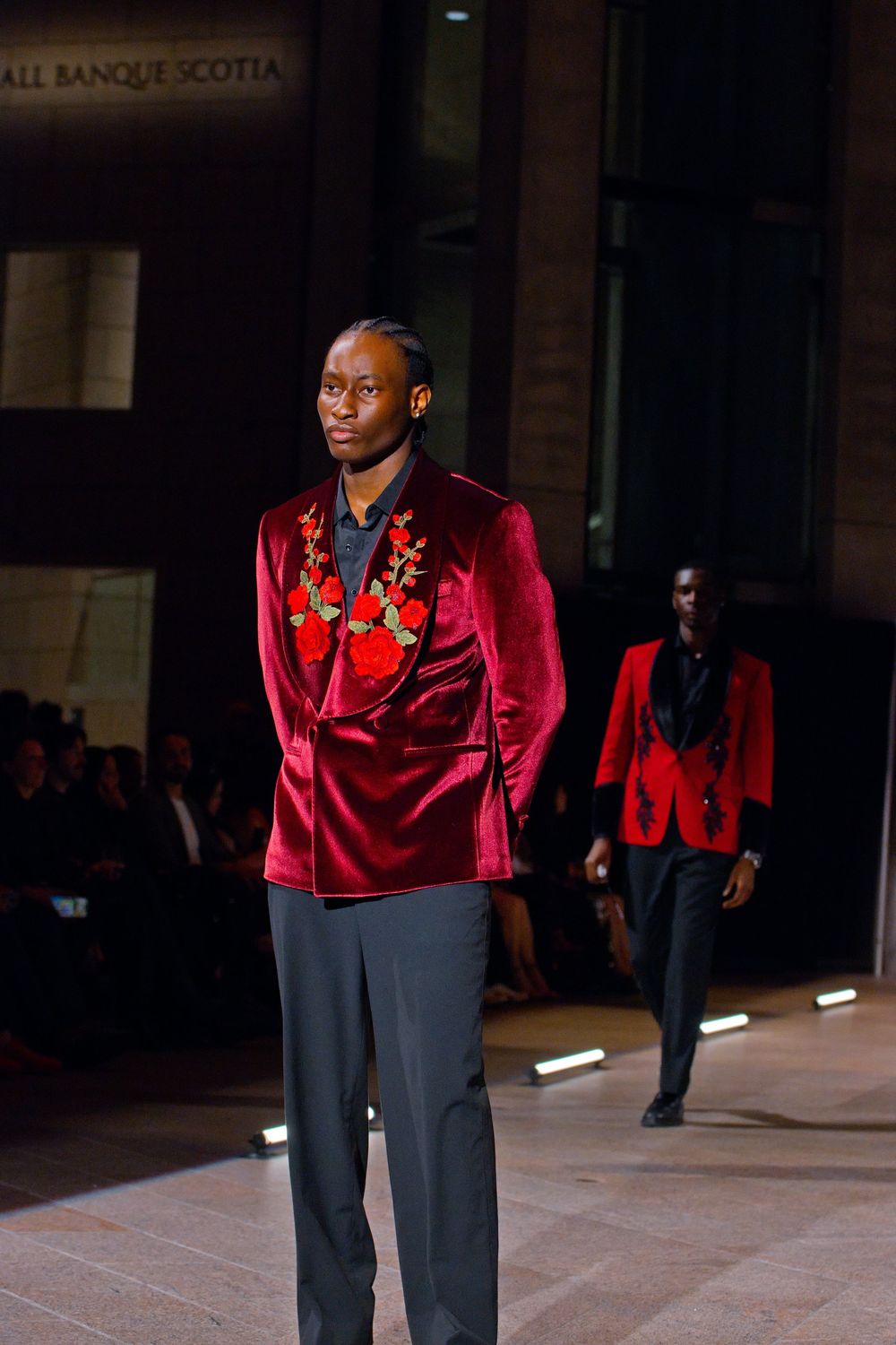 Male model wears red velvet blazer with floral embroidery on runway at formal fashion show event.