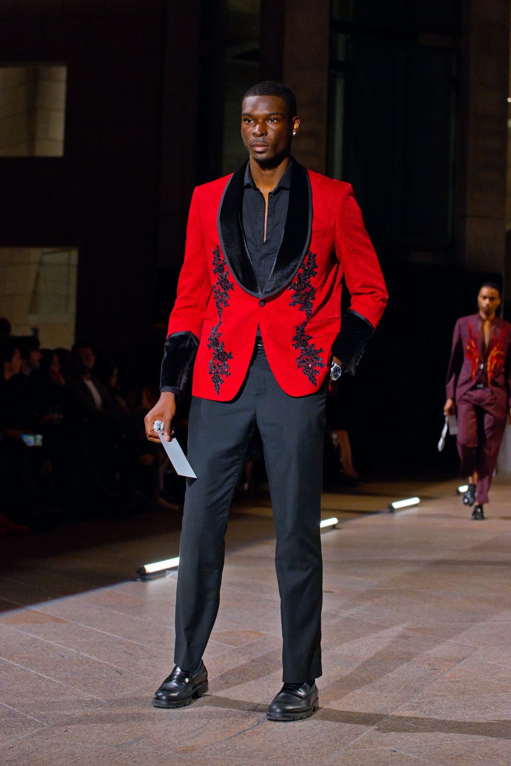 Male model walks runway in red velvet blazer with black floral embroidery and black trousers.