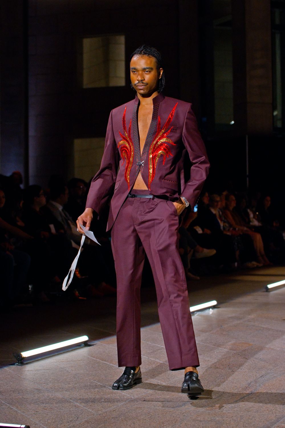 Male model walks runway in burgundy suit with red flame embroidery and open chest at fashion show.