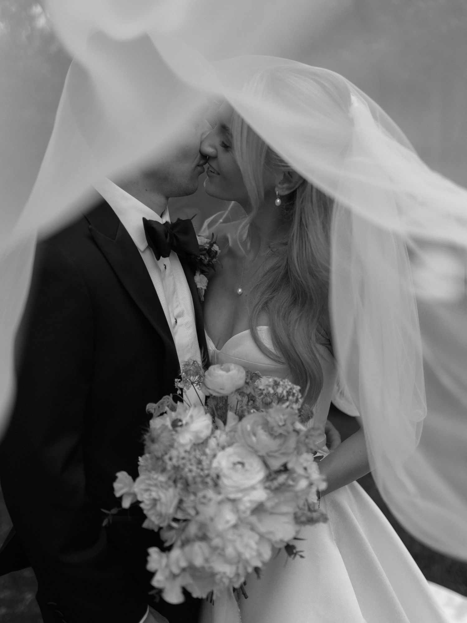 Black and white wedding photo of bride and groom kissing under flowing veil, bride holding floral bouquet.