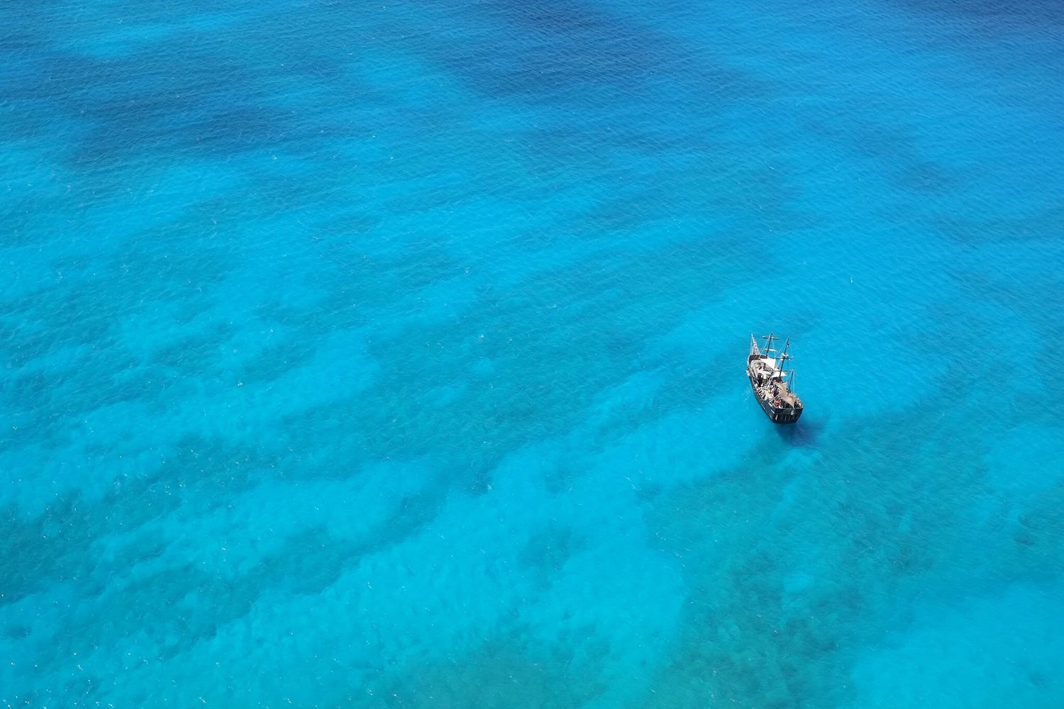 Aerial view of a single boat floating on crystal clear turquoise ocean water with visible coral reef below.