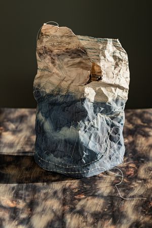 Sculptural crumpled fabric vessel in dark blue and white tones, stitched together and placed on a textured dark surface.