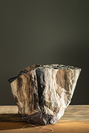Crumpled fabric vessel with earth tone marble pattern and white wire stitching on sandy surface.
