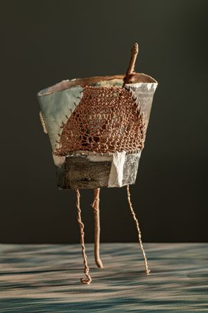 Handcrafted ceramic and copper wire mesh figurine with twig arms and wire legs on wooden surface.