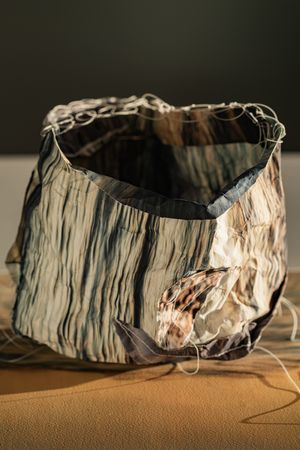 Open-top crumpled fabric bowl with striped pattern, white wire rim, and shell detail on sand.