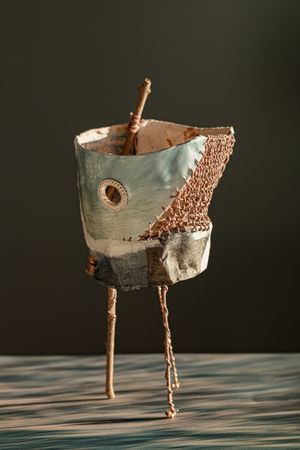 Whimsical sculptural figure made from fabric and mixed materials, standing on stick legs with copper wire details on a wooden surface.