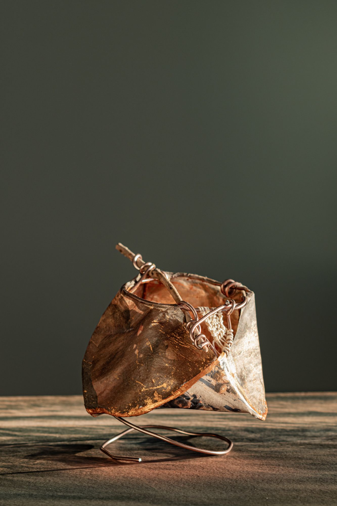 Geometric copper and silver sculptural form balanced on a wire stand, with intricate metallic details on a wooden surface.