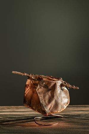 Abstract copper-toned sculptural vessel with stick handles, resting on a curved metal stand on a wooden surface.