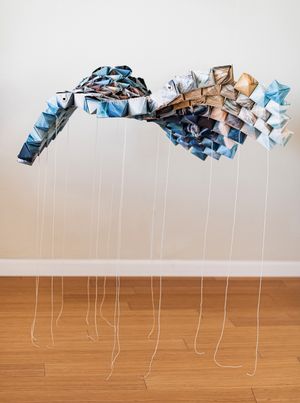 Suspended origami-like sculpture in blue and tan tones with hanging strings above wooden floor.