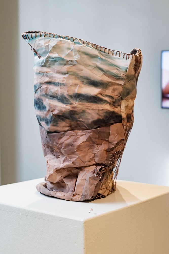 Crumpled paper bag sculpture displayed on white pedestal in art gallery, featuring textured brown tones and stitched edges.