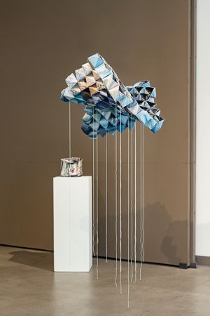 A geometric blue origami sculpture suspended mid-air with white strings above a white pedestal in a modern gallery.