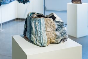A crumpled fabric sculpture with blue and beige tones displayed on a white pedestal in a gallery.