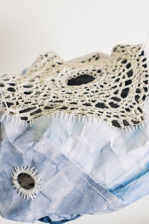 Close-up of a crumpled blue fabric art piece with intricate cream crochet lace trim and embroidered eye detail.