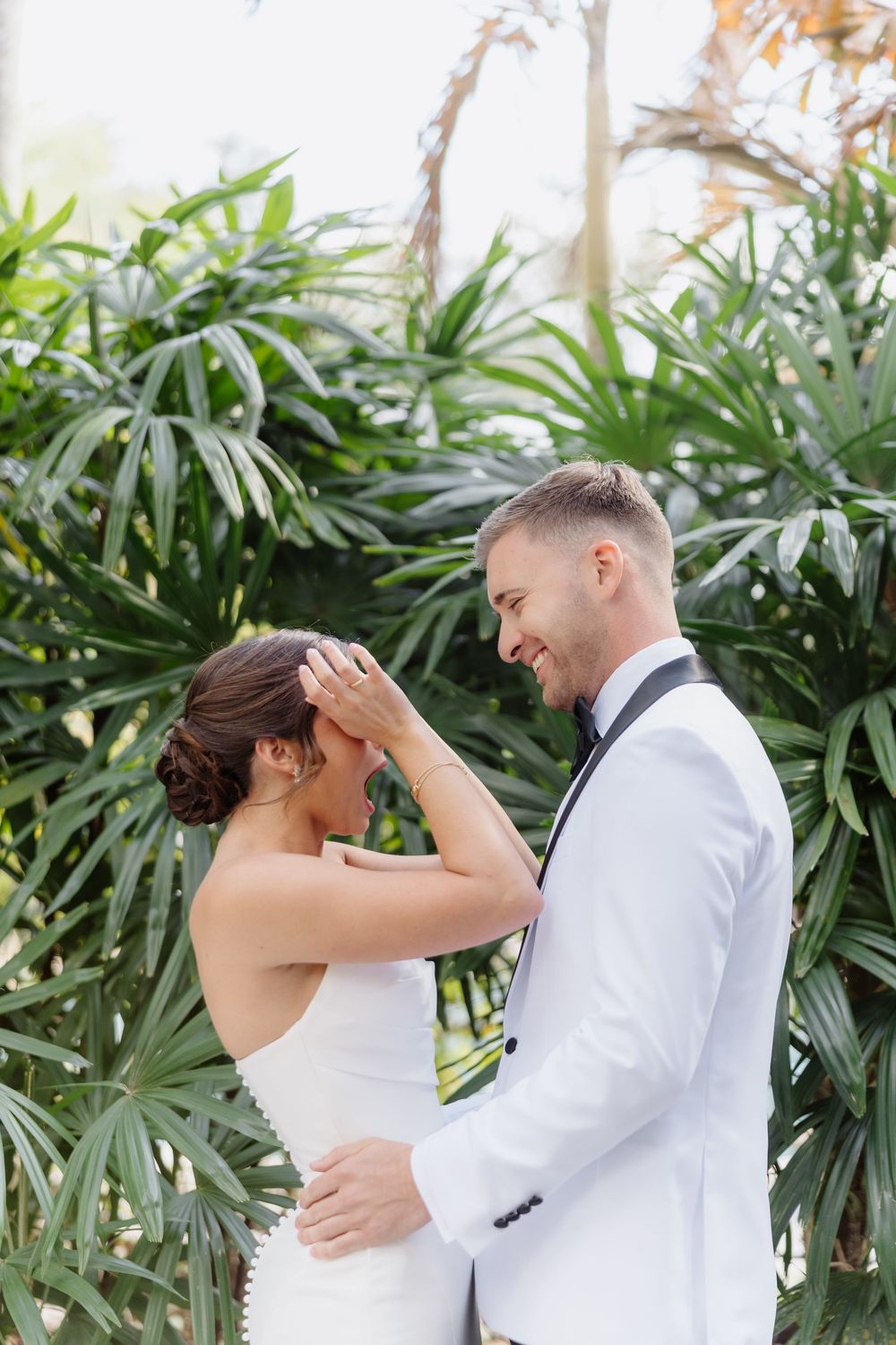 Smiling groom in white tuxedo tenderly holds bride's face amid lush tropical greenery.