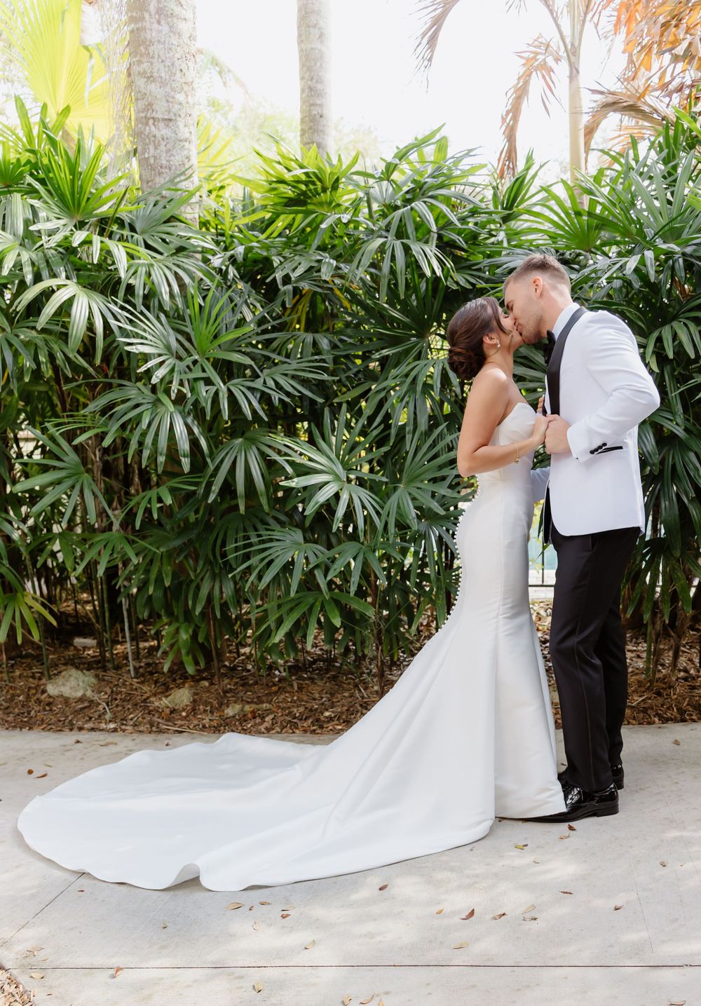 Bride and groom share a romantic kiss in front of tropical palm plants, bride in fitted mermaid gown with train.