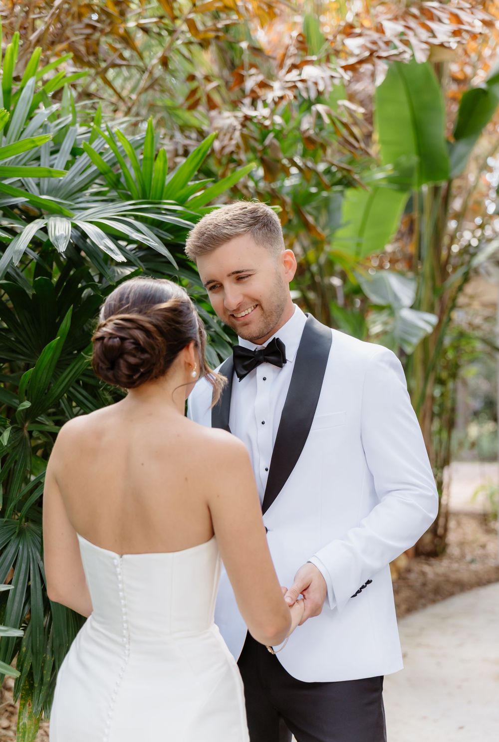 Groom in white tuxedo jacket smiles at bride during first look surrounded by tropical greenery.