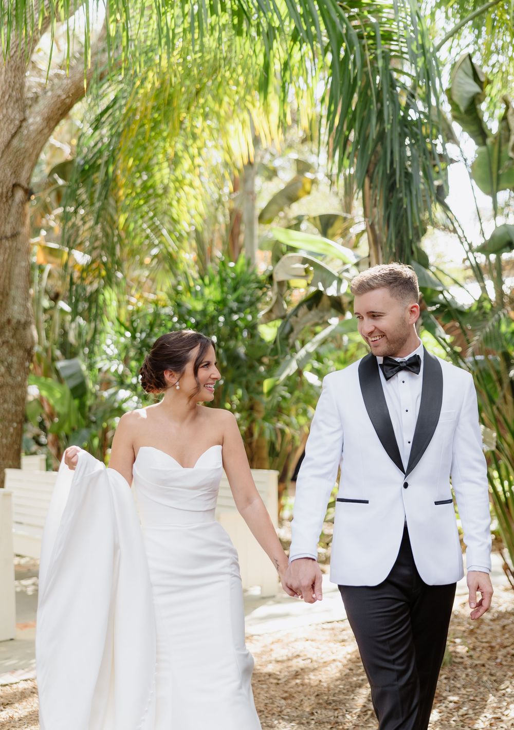 Bride and groom walk hand in hand smiling among tropical palm trees in white tuxedo and gown.