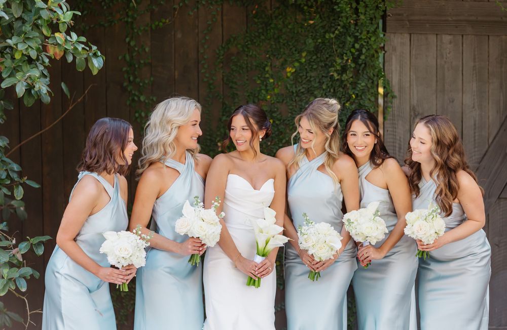 Bride in strapless gown stands with five bridesmaids in matching light blue halter dresses holding white bouquets.