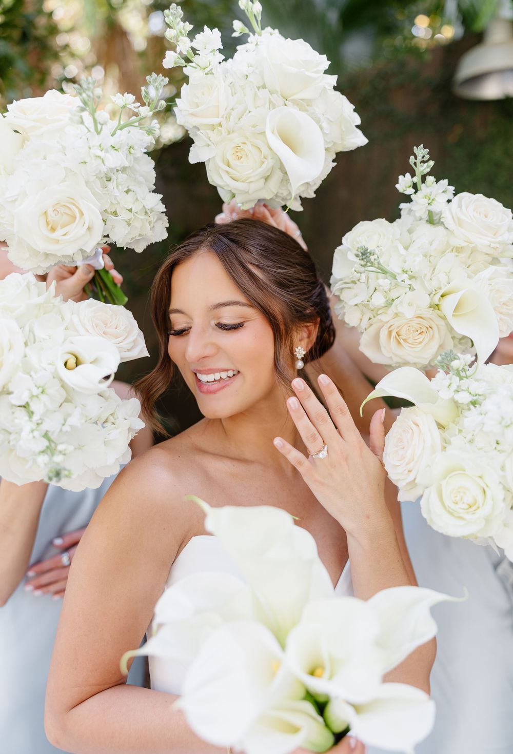 Bride surrounded by white rose and calla lily bouquets held by bridesmaids in soft light.
