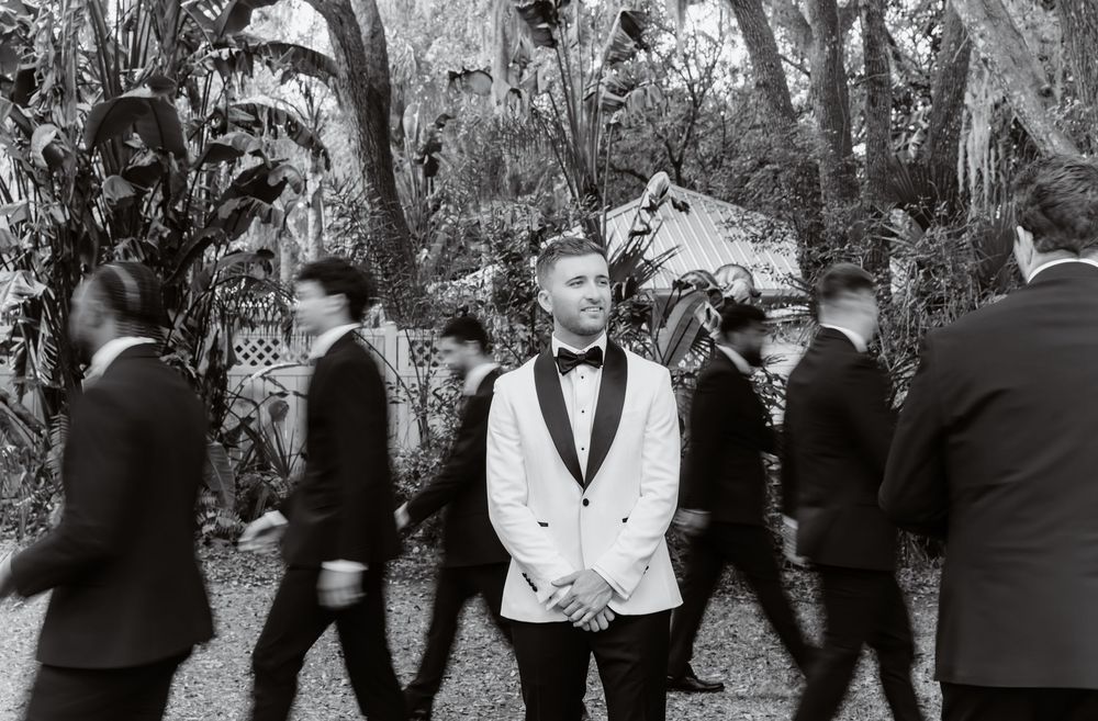 Groom in white tuxedo stands still among blurred groomsmen in black suits in a lush garden setting.
